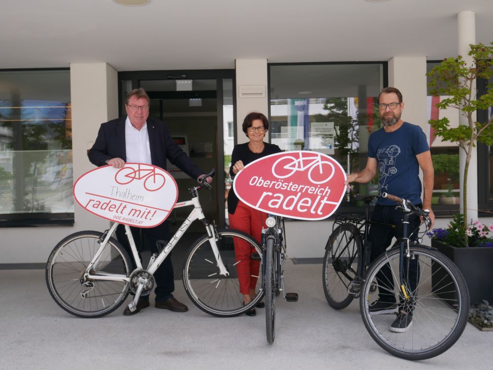 Schilder mit "Thalheim radelt mit" und "Oberösterreich radelt mit" werden von drei Menschen auf Rädern vor einem Gebäude gehalten